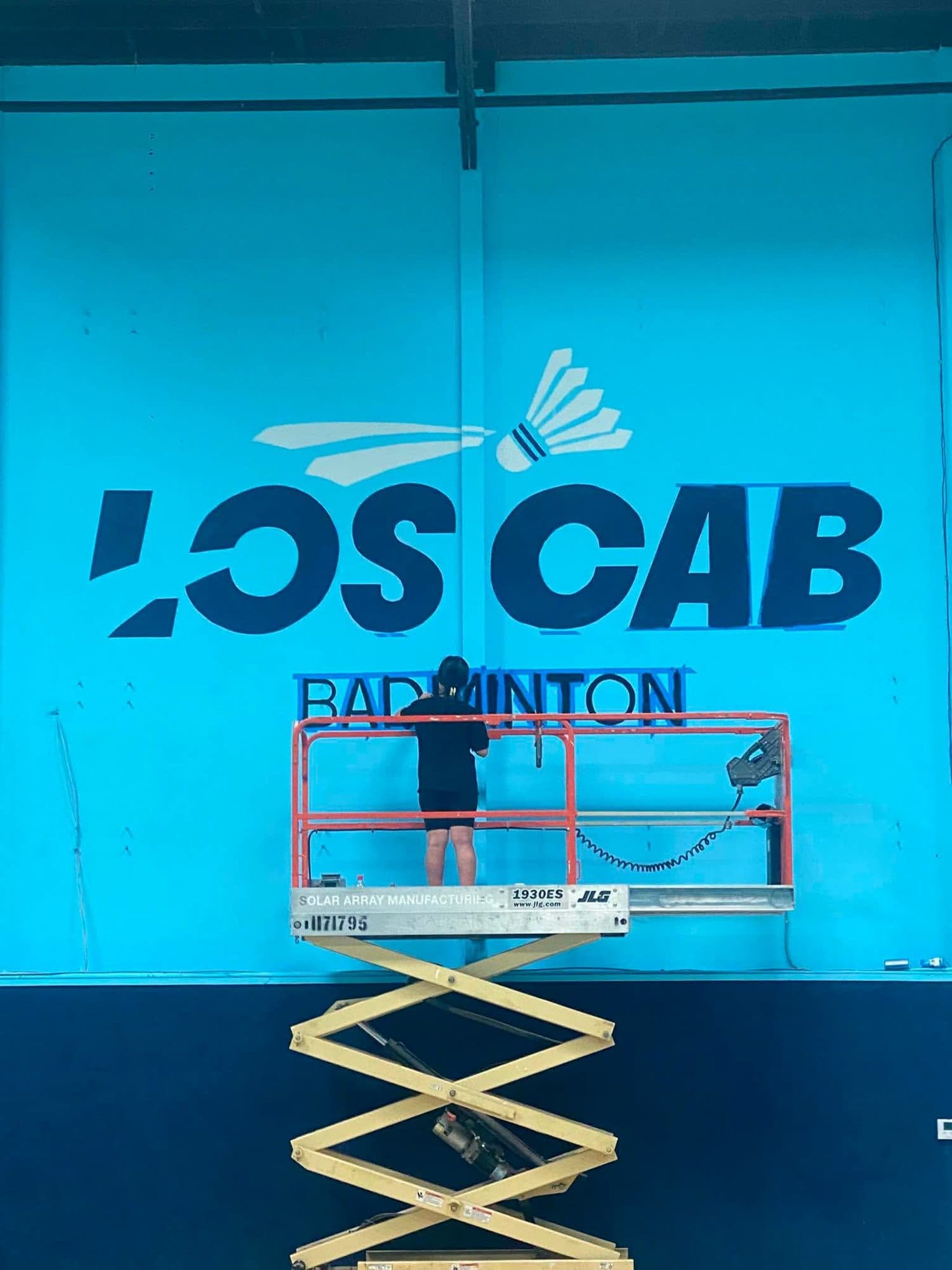 Eve painting the LOS CAB Badminton logo on a tall blue wall from a scissor lift